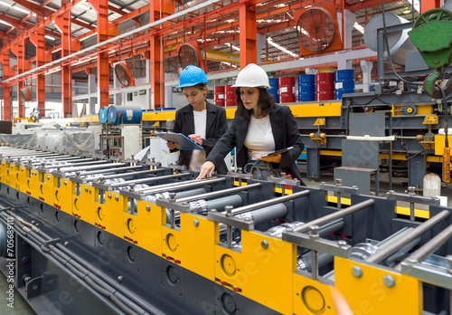 Two women operate machinery in a factory, wearing safety gear and focused on their task.
