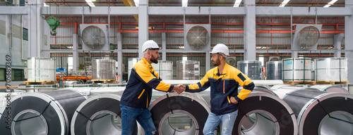 Two people shaking hands in front of large rolls of steel at an industrial facility.