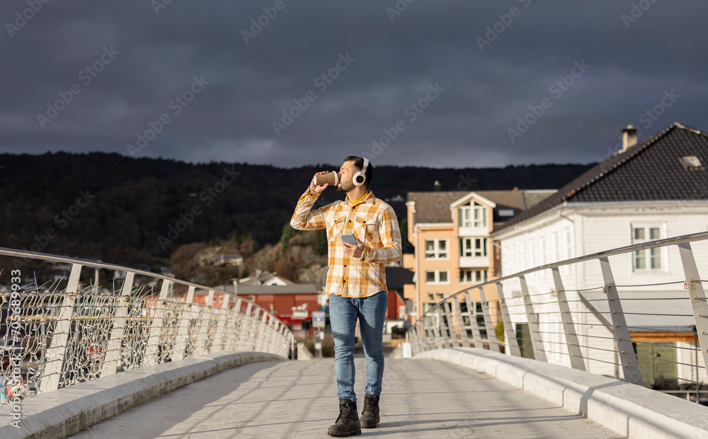 Hispanic man walking towards camera drinking coffee and listening to music with his headphones with the city in the background Hispanic man walking towards camera drinking coffee and listening to music with his headphones with the city in the background