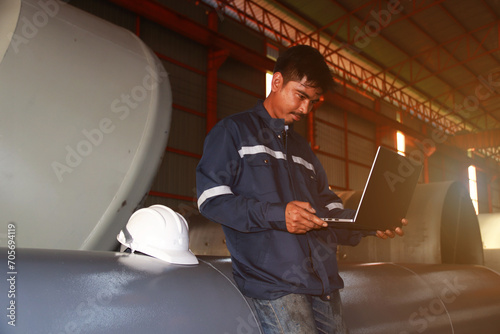 Chief engineer in hard hat walks through modern factory lighting while holding laptop. Successful handsome man in modern industrial environment.