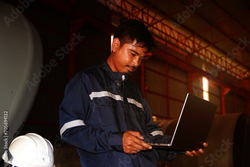 Chief engineer in hard hat walks through modern factory lighting while holding laptop. Successful handsome man in modern industrial environment.