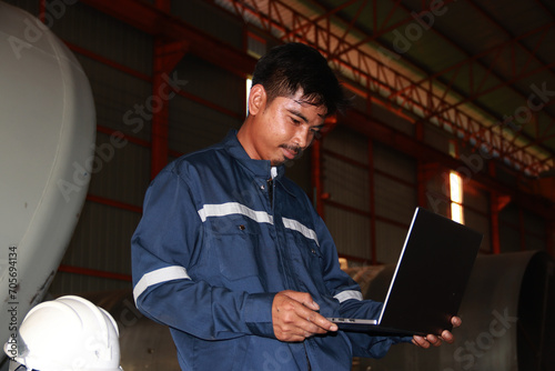 Chief engineer in hard hat walks through modern factory lighting while holding laptop. Successful handsome man in modern industrial environment.