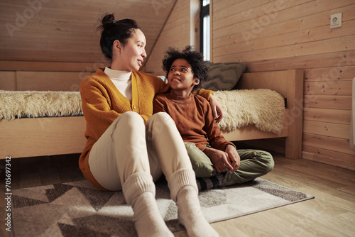 Young woman in warm casualwear sitting on the floor next to her cute son and talking to him while spending weekend or vacation in cottage photo