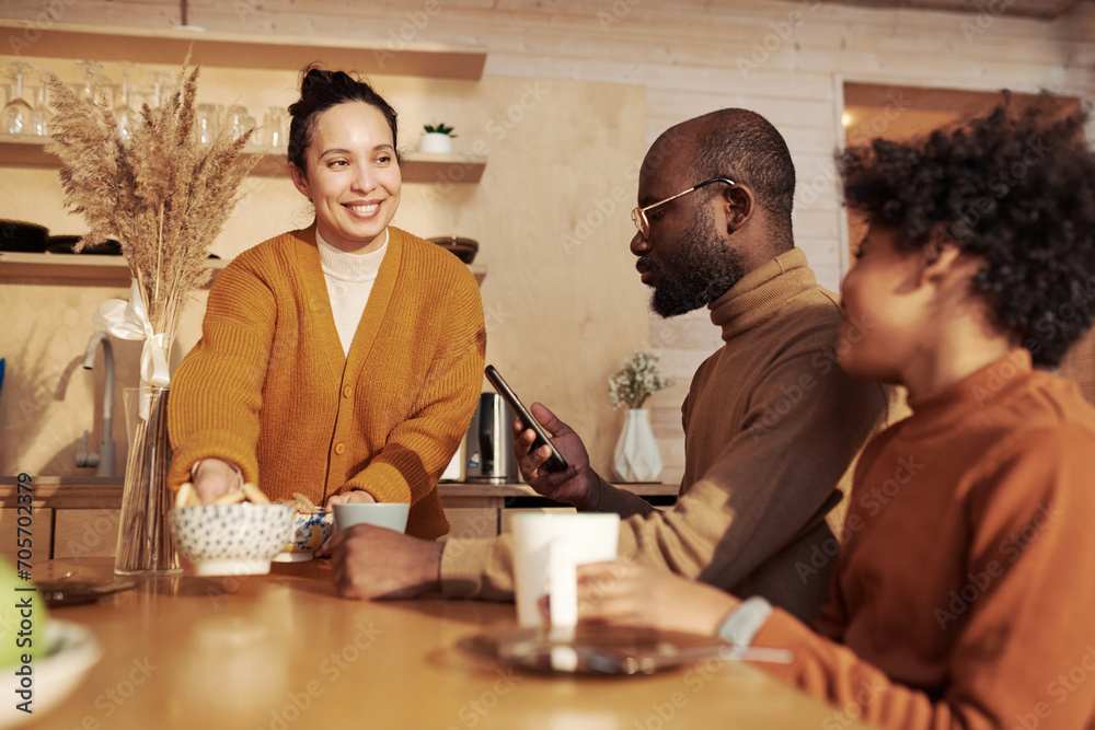 Happy young woman putting two porcelain bowls with muesli on wooden table and looking at her son while serious man texting in smartphone Happy young woman putting two porcelain bowls with muesli on wooden table and looking at her son while serious man texting in smartphone