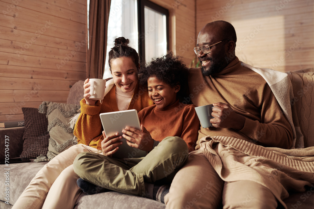 Happy young intercultural family of three looking at screen of tablet held by cute boy while sitting on couch and watching sitcom at leisure - obrazy, fototapety, plakaty 