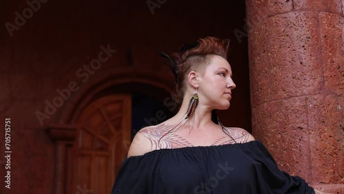 Charming woman with tattoos on her body, original feather decorations smiles in ancient building. 