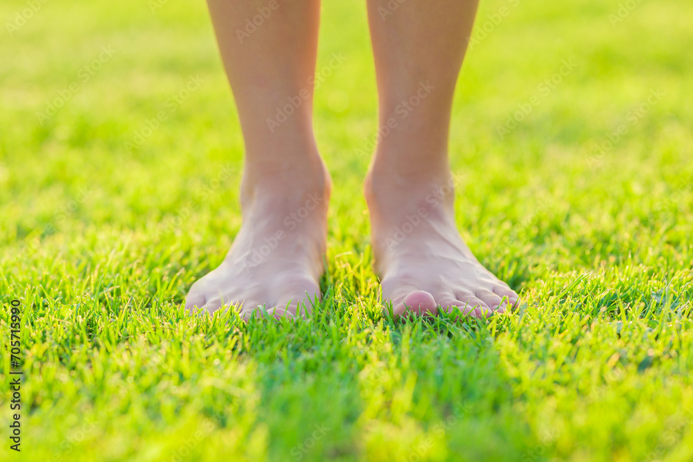 Young adult woman barefoot standing on fresh green grass at park in beautiful warm sunny summer day. Front view. Closeup. Healthy walk.