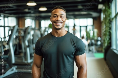 Attractive muscular black man in sportswear stands against the backdrop of a gym and exercise equipment. Personal trainer in a sports club smiles and looks at the camera.