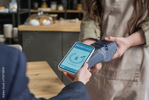 Hands of young waitress holding payment terminal while female guest of cafe holding smartphone with online banking operation data on screen photo