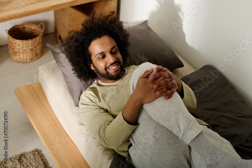 Smiling man in cotton pullover and sweatpants doing stretching exercise for legs after sleep while lying on bed in the morning photo