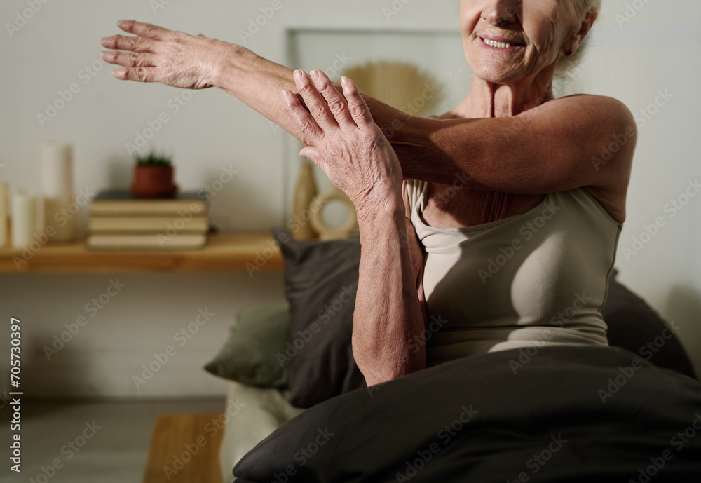 Cropped shot of senior woman in cotton tanktop sitting on bed after sleep and stretching arm aside while doing physical exercise