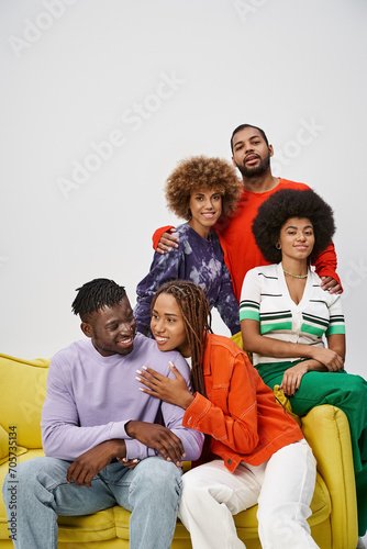 happy african american friends in bright casual attire sitting together on yellow couch on grey