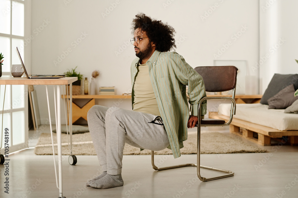 Multiethnic young man in casualwear doing squats in front of laptop while holding by chair and looking at screen during physical exercise