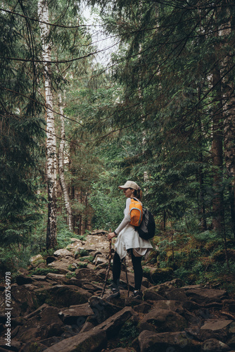 A girl traveler, with a backpack and trekking poles, climbs a rocky path among a green forest