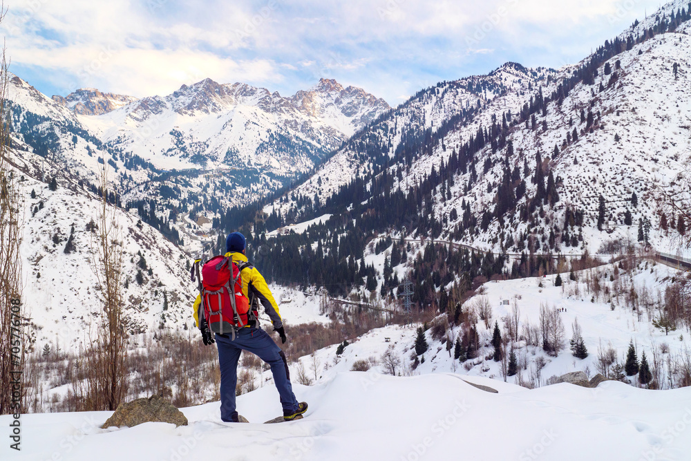 A male tourist with a backpack looks at the mountain panorama in winter. A guy with a red backpack in a yellow jacket. Mountain tourism in winter