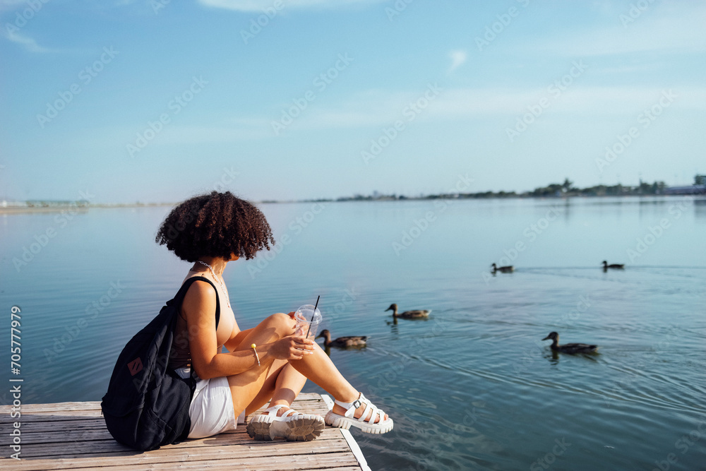 Beautiful mixed race female teenager sits on wooden pier and drinks cocktail in plactic glass. Charming african girl in casual clothes and with black backpack smiles sincerely near water. Sunny day.