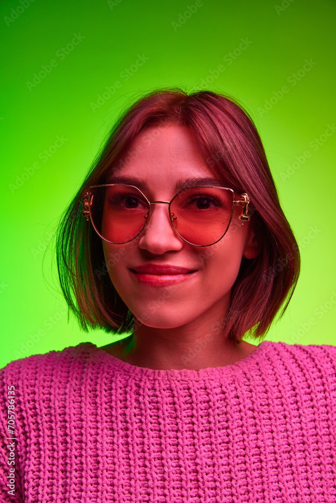 Fototapeta premium Vertical portrait of young beautiful woman in modern, fashion sunglasses looking at camera against green background in pink neon light, filter. Concept of art, beauty, youth, human emotions, style. Ad