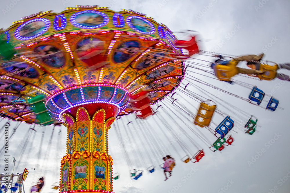 Detalhe de um brinquedo gigante de rotação no parque de diversões com poucas pessoas, em um dia nublado. Foto feita de baixo para cima. - obrazy, fototapety, plakaty 