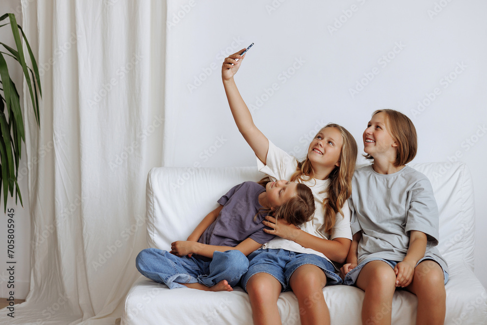 Three girls take a selfie while sitting on the sofa. Three sisters smiling in casual clothes against a white wall. Concept for advertising a mobile application or content for the whole family.