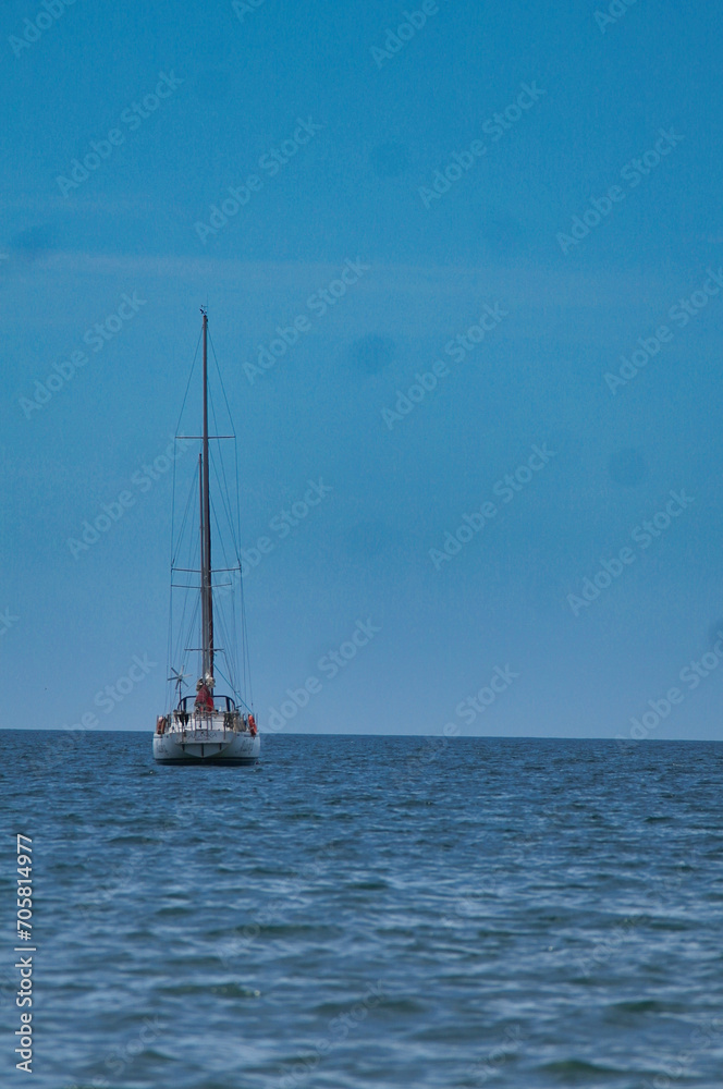 Fototapeta premium The image depicts a sailboat on the open sea under a clear blue sky. The main subject of the image is a white sailboat with a tall mast, floating on the calm sea. The sea has gentle waves and reflects