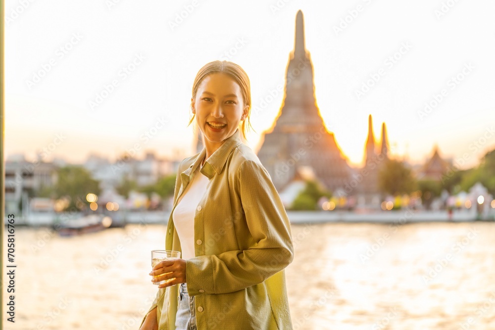 Young Asian Woman Traveler Holding a Drink While Enjoying The Sunset Moments of Wat Arun by the Chao Phraya Riverbank in Bangkok, Thailand