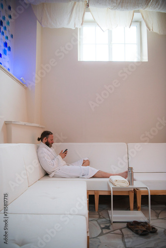 Man with beard and hair pulled back to ponytail relaxing in a plush white robe on a white couch using smart phone