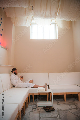 Man with beard and hair pulled back to ponytail relaxing in a plush white robe on a white couch using smart phone