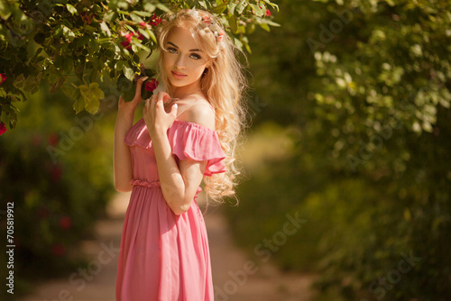 Beautiful young woman with long curly hair and perfect skin wearing pink linen dress posing near blooming roses in a garden. Nude make up. Close up portrait © olenakucher