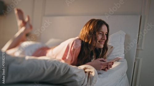 Young woman using mobile phone at home. Smiling happy female lying in bed typing