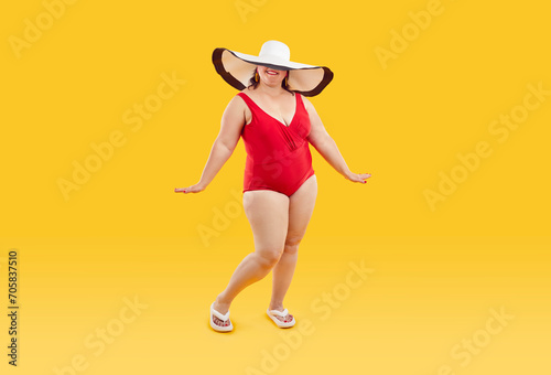 Full length studio portrait of happy beautiful young fat plump woman wearing red one piece swimsuit and big wide holiday beach sun hat that covers her eyes posing isolated on yellow color background