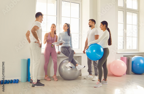Group of young sporty people talking with coach, fitness club sportswomen holding group discussion, team standing with gym balls. Active friends before, after exercise or sport instruction together