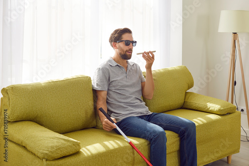 Quadro em tela Portrait of a young bearded blind man in black eyeglasses holding cane and using digital assistant on mobile phone sitting on sofa at home
