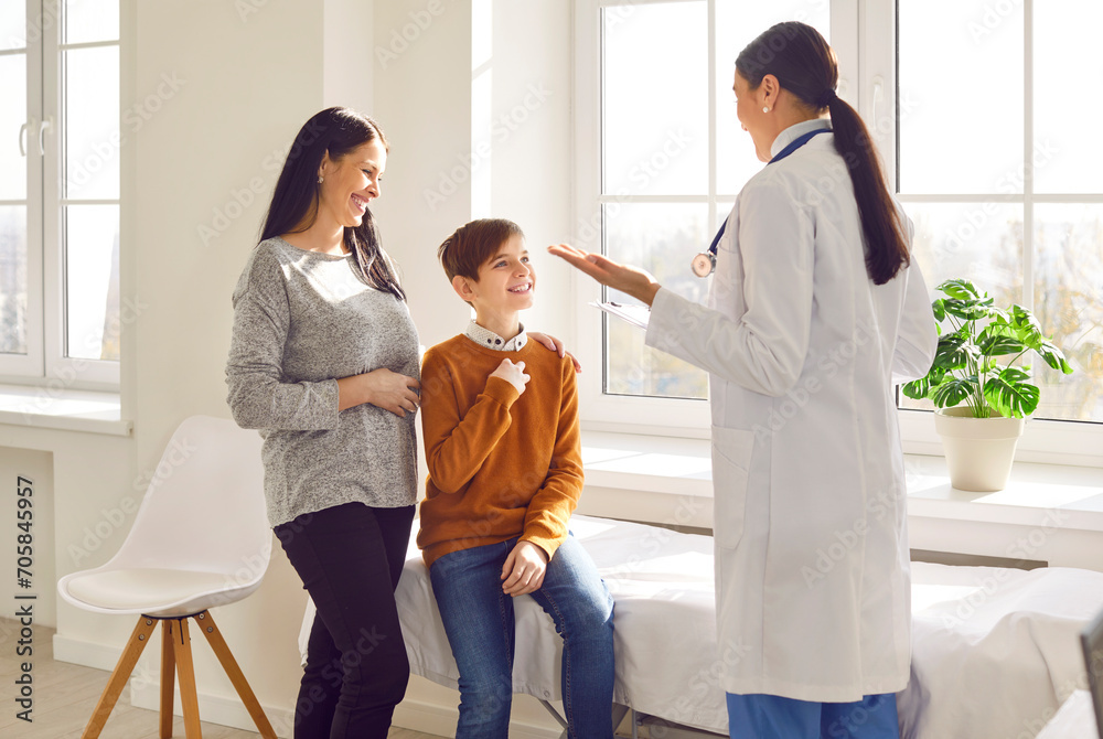 Woman pediatrician examining preteen boy patient who came with his mother. Cheerful doctor talking to mother and son at pediatric check up appointment in medical clinic. Children medical care concept