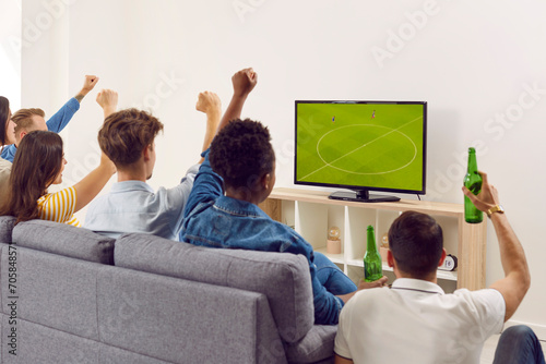 Group of happy diverse friends watching soccer on TV. Young multiracial people sitting on sofa, looking at television screen, watching football, drinking beer, raising hands up and cheering. Back view