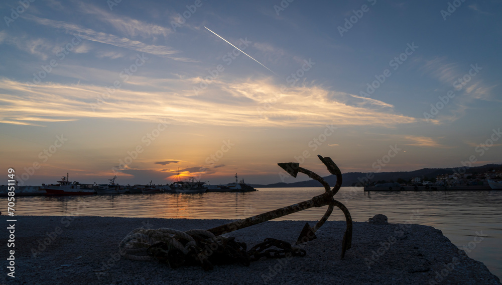 An old rusty anchor with a rope lies on the edge of a pier in a bay with fishing boats in Skioni, Greece