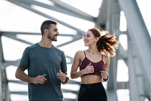 Young adult couple running together in the city looking at each other while jogging. Happy athletic male and female in sportswear training workout outside. © carlesiturbe