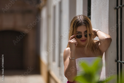 South American woman, young, beautiful and blonde with white top, pink pants and sunglasses, posing leaning on a white wall, in a sensual and provocative attitude. Concept trend, beauty, fashion. © Manuel