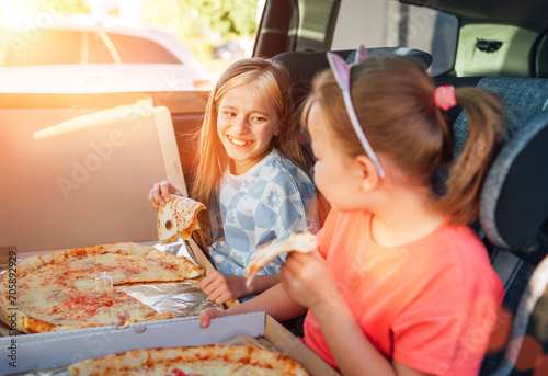 Portrait of two positive smiling sisters eating just cooked italian pizza sitting in child car seats on car back seat. Happy childhood, fastfood eating or auto jorney lunch break concept image. photo
