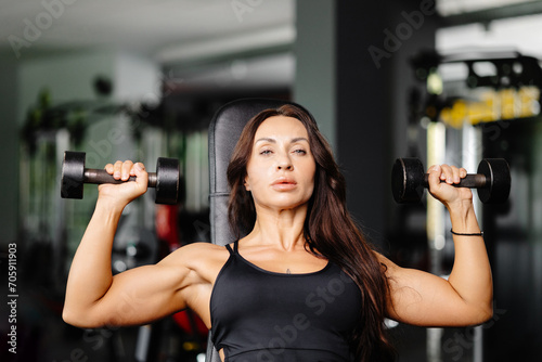 Young fit woman practicing dumbbell press while sitting on a bench. Slim female in sportswear exercises with weights in a gym