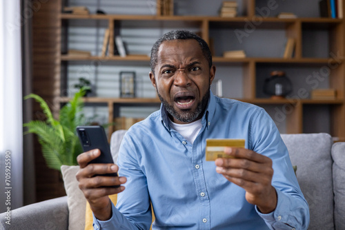 Close-up portrait of African American man sitting on sofa at home, holding credit card and phone, angry and worried looking at camera. photo