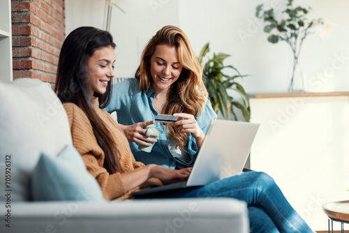 Two happy young women using computer while paying with credit card their vacation sitting on the couch at home. photo