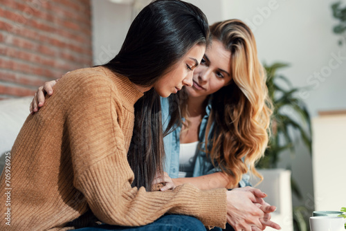 Pretty young woman supporting and comforting her sad friend while sitting on the sofa at home. photo