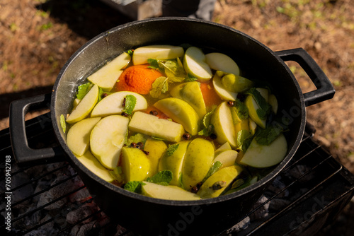 Steaming white mulled wine in a pot with sliced apples, mandarins, and fresh mint on a grill, ready for a cozy outdoor gathering