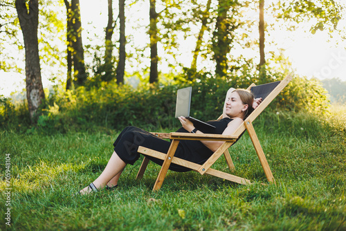 A cute pregnant woman is sitting in a park and working remotely on a laptop. Online work on maternity leave. Modern pregnant woman © Дмитрий Ткачук