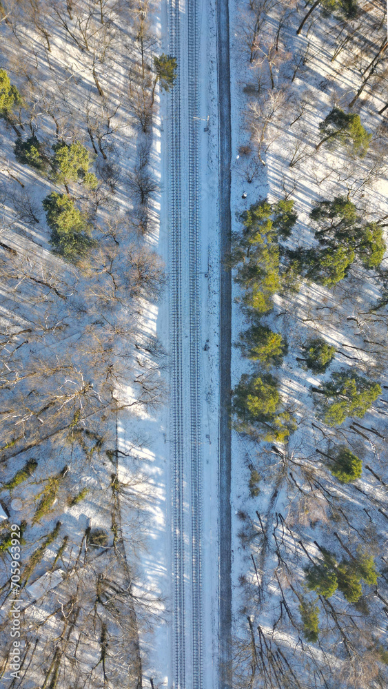Railway tracks through the forest in snow, empty railway traction, concept of train delays due to winter weather conditions, vertical, aerial view. Railway tracks through the forest in snow, empty railway traction, concept of train delays due to winter weather conditions, vertical, aerial view.