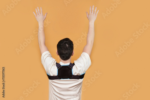A young guy wears a special posture corrector to keep his back straight and healthy and does special exercises with arms up, rear view. The boy has a corset on his back, back hurts photo