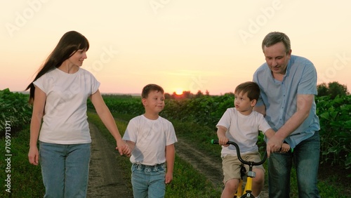 Parent looks at child while father aids son riding bicycle against late sunset. Son pedals bicycle with father help while sibling walks hand in hand with mother on ground road. Summer vocation