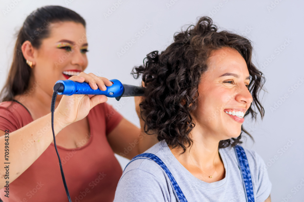 Young Latina women are enjoying a moment of laughter while one of them is working on the other's hair; both are wearing big smiles