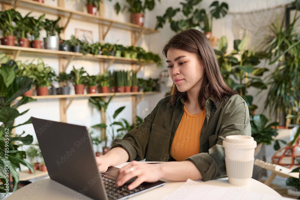 Young female entrepreneur working on her laptop sitting at table in plant store Young female entrepreneur working on her laptop sitting at table in plant store