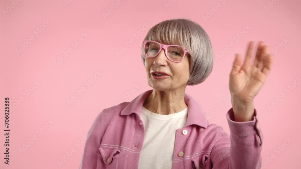 Stylish grandmother greets and waves hand warmly. She is in a pink blouse and pink glasses against a pink background. The woman is fashionably dressed in a single color, emphasizing a modern style. Stylish grandmother greets and waves hand warmly. She is in a pink blouse and pink glasses against a pink background. The woman is fashionably dressed in a single color, emphasizing a modern style.